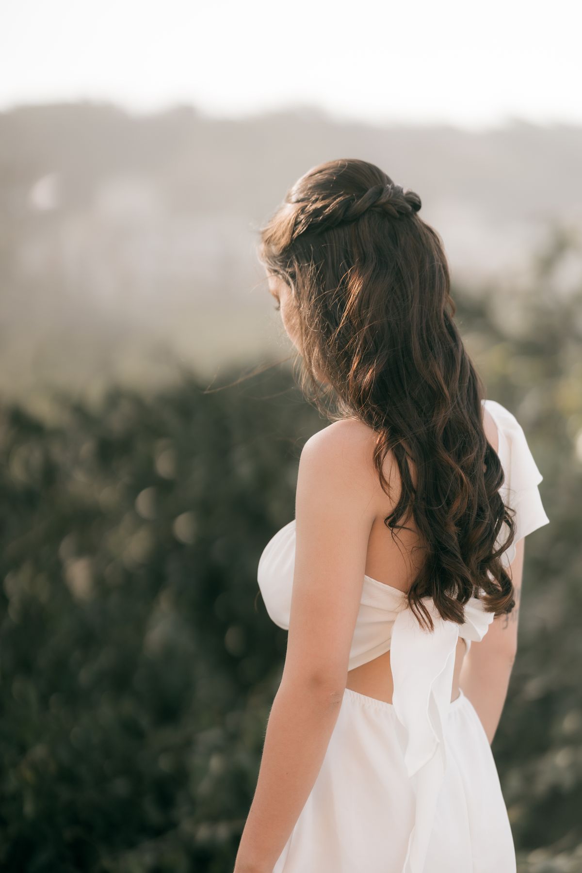 Woman seen from behind with long, dark brown curled waves half up in a knot at the crown, wearing a white dress with a bow at the back against blurred hills