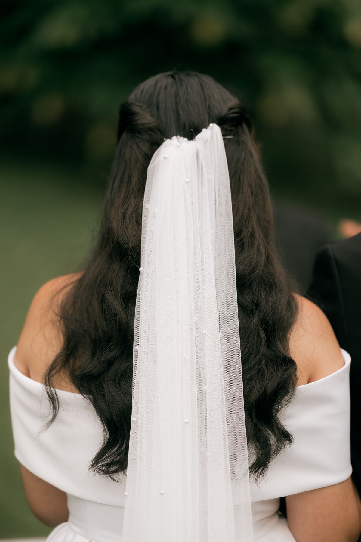 Woman seen from behind with long dark wavy hair half up, wearing a long white veil with tiny pearls and a white off the shoulder dress, outdoors