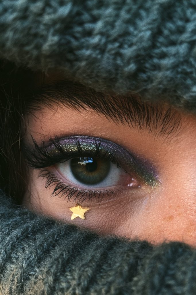 Close up of an eye with purple and green witchy eyeshadow, thick lashes and a tiny gold star under the eye