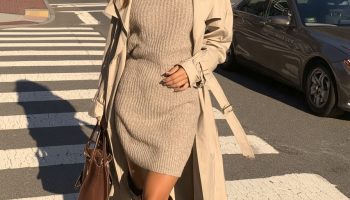 Woman in dark brown knee high boots with an oatmeal sweater dress and a beige trench coat crossing a street