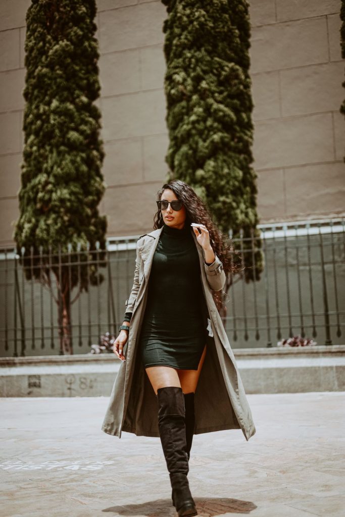 Woman in sunglasses and a long grey-beige coat over a black high-neck dress with tall dark over-the-knee boots