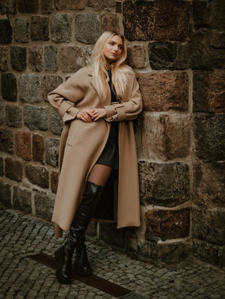 Woman in a long camel coat and black knee-high boots leaning on a stone wall