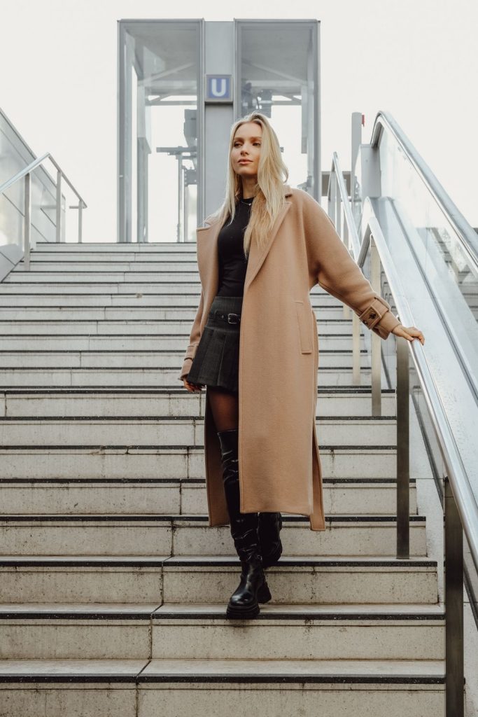 Woman walking down stairs in a long tan coat, a black outfit and black boots