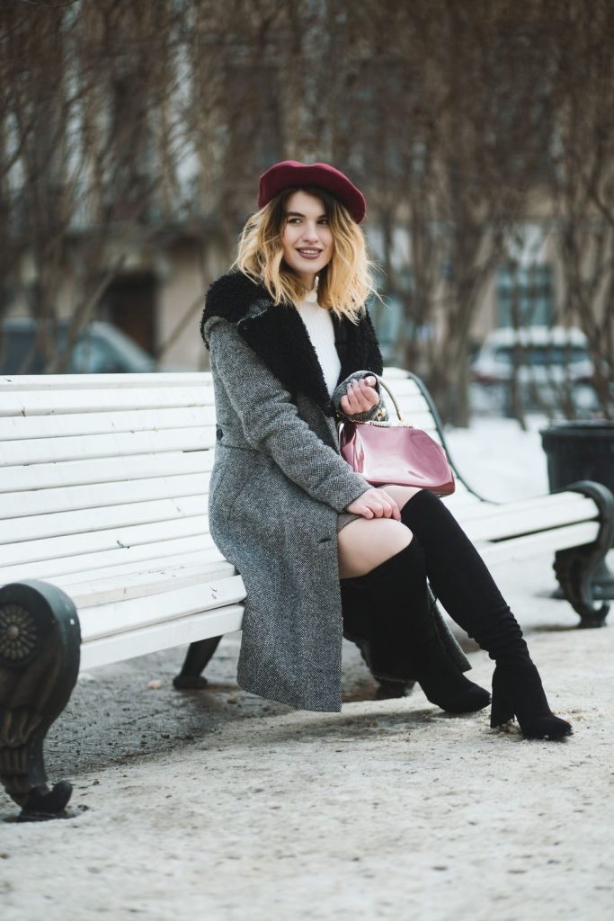 Woman in a maroon beret and a grey tweed coat with a dark collar sitting on a white bench with a pink handbag and black knee-high boots