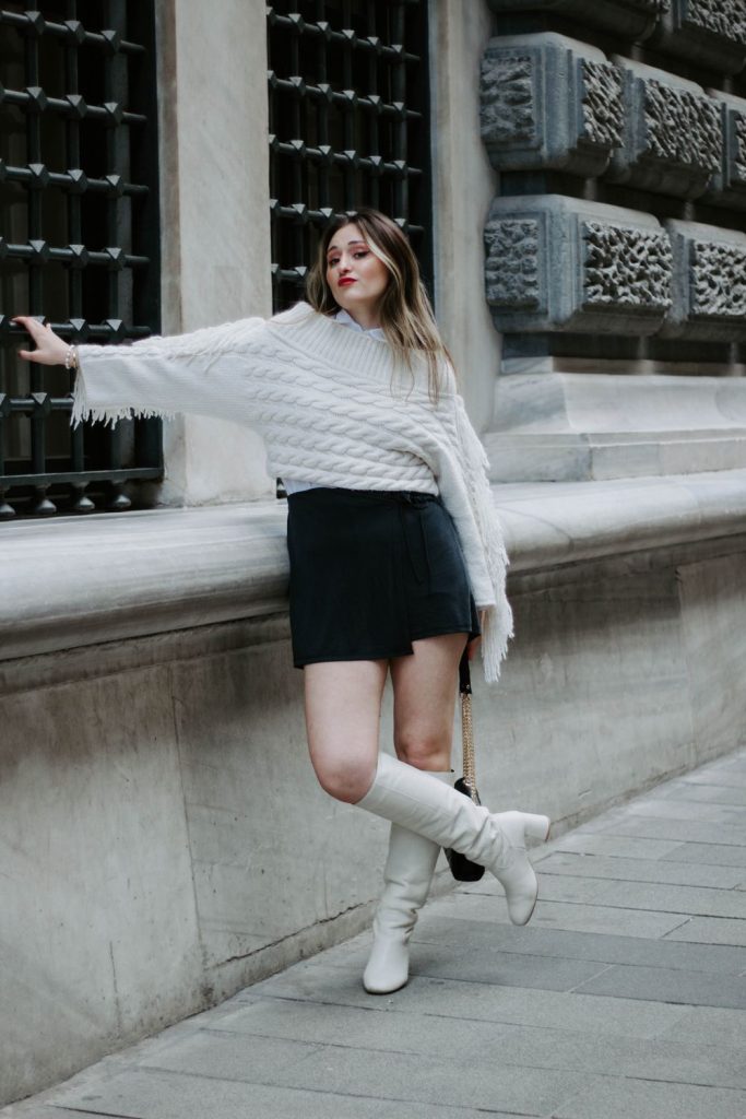 Woman in a white fringed knit sweater and a black mini skirt with white knee-high boots leaning against a window