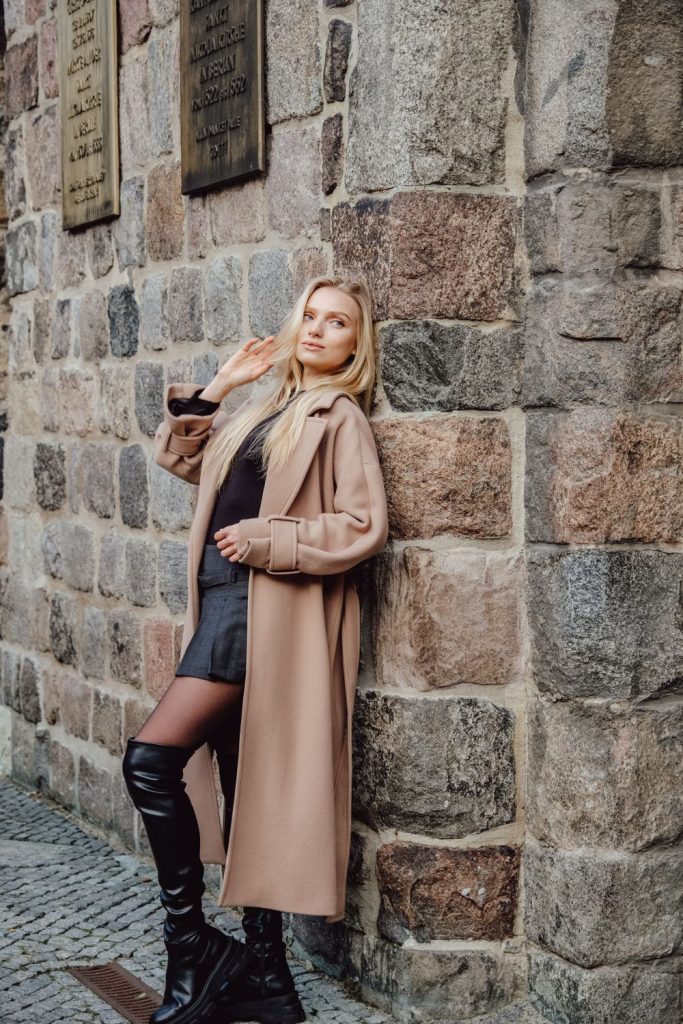 Woman in a long camel coat, a black dress and black tights with knee-high boots leaning on a stone wall