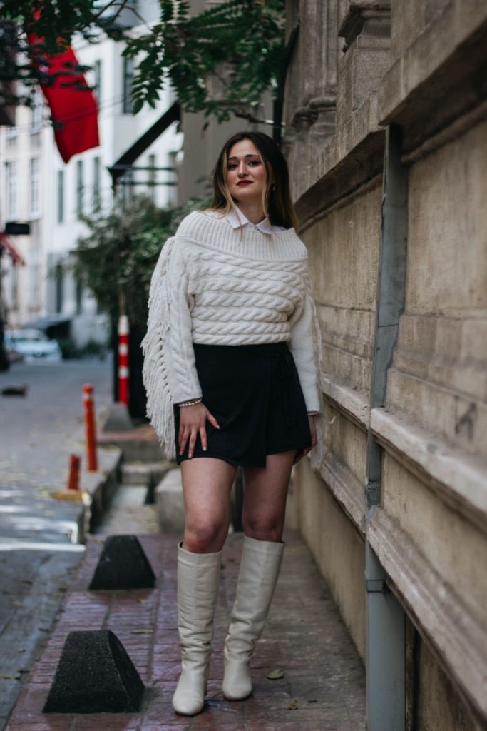 Woman in a white cable knit sweater with a collared shirt, a black skirt and white knee-high boots on a city street