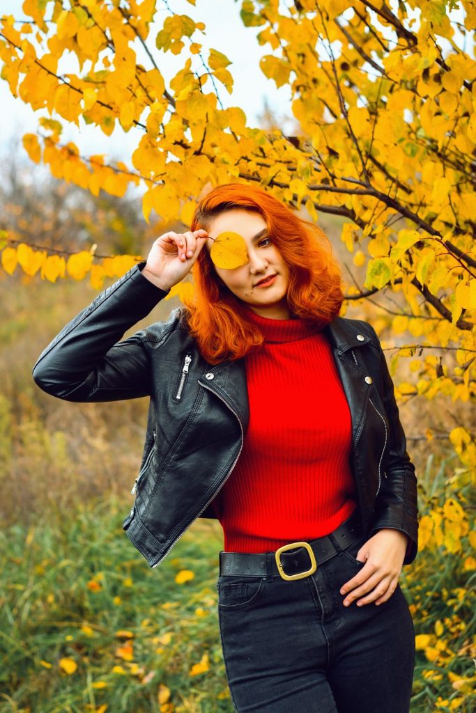 Woman with red-orange hair in a fitted black leather biker jacket over a red top with a black belt with a gold buckle holding a yellow leaf