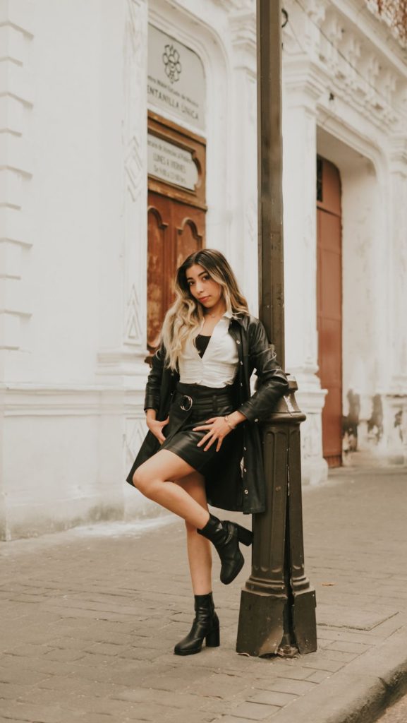 Woman leaning on a lamppost in a black leather jacket, a white top, a black leather skirt and black ankle boots