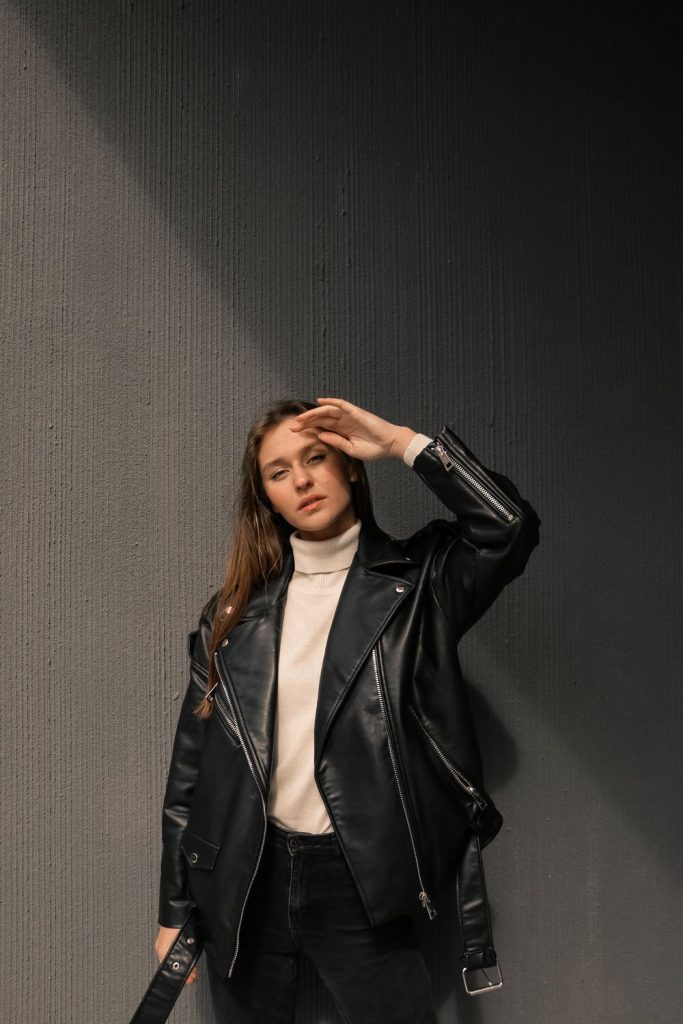Woman with long hair in an oversized black leather jacket over a white turtleneck standing against a grey wall
