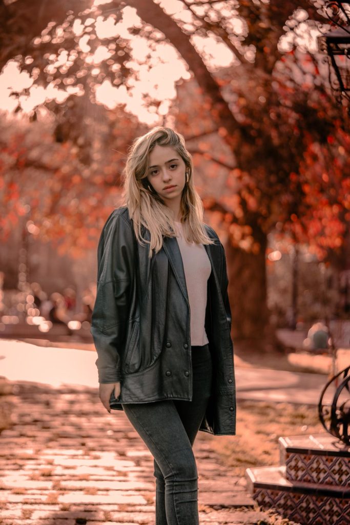 Woman with long blond hair in an oversized dark leather jacket over a blush top and grey trousers in an autumn park