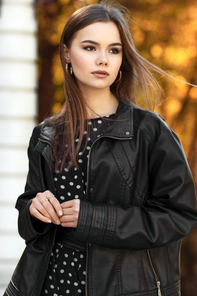Woman with long brown hair and hoop earrings in an oversized black leather jacket over a black and white polka dot dress