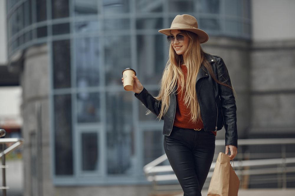 Woman in a tan fedora, sunglasses, a black leather jacket, a rust top and black trousers holding a coffee and a shopping bag