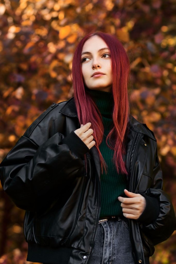 Woman with long burgundy hair in an oversized black leather jacket over a dark green turtleneck among autumn leaves