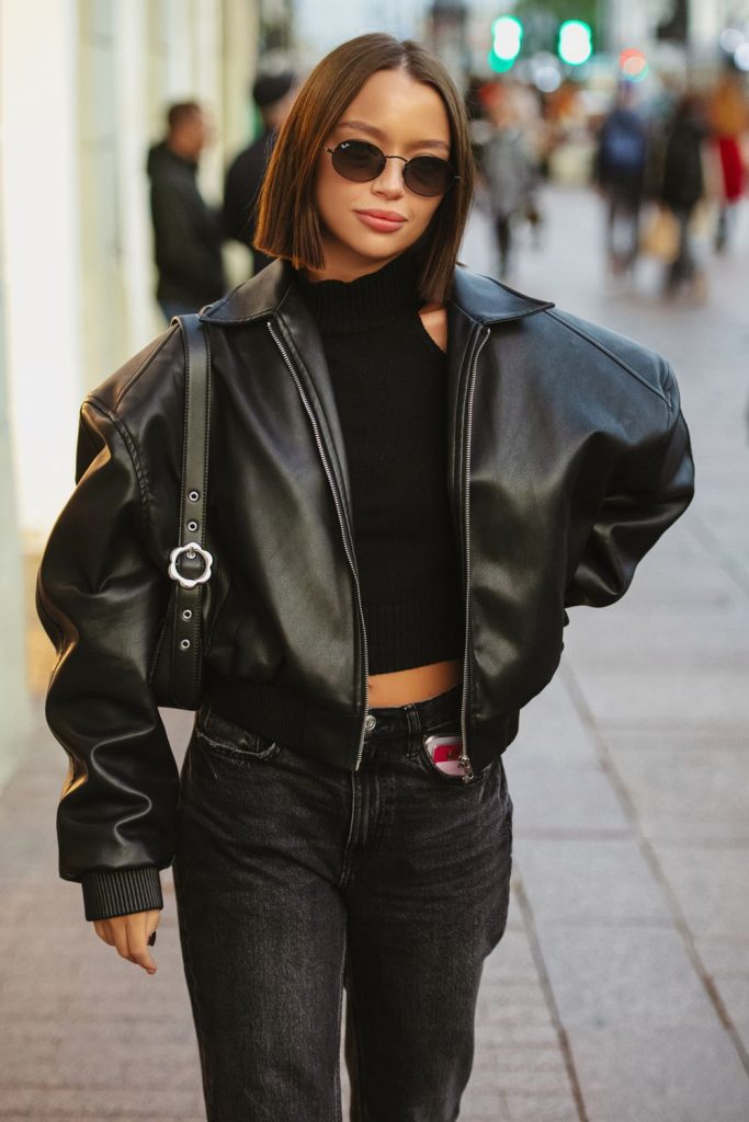 Woman in sunglasses and an oversized black leather jacket over a black top with black jeans and a black shoulder bag on a city street