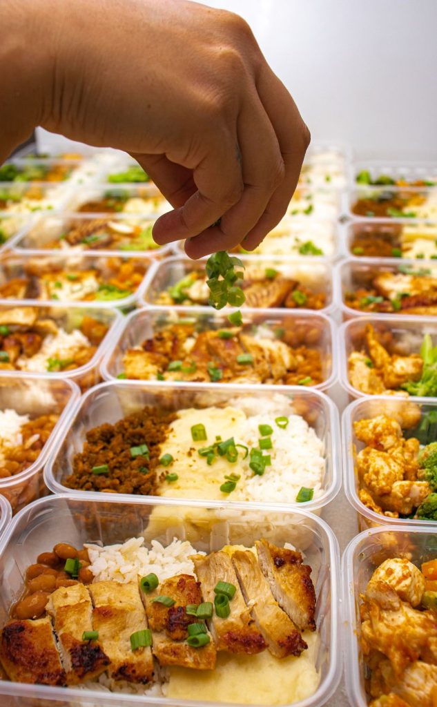 Hand garnishing rows of takeaway containers filled with rice, chicken and sides, packed to send home