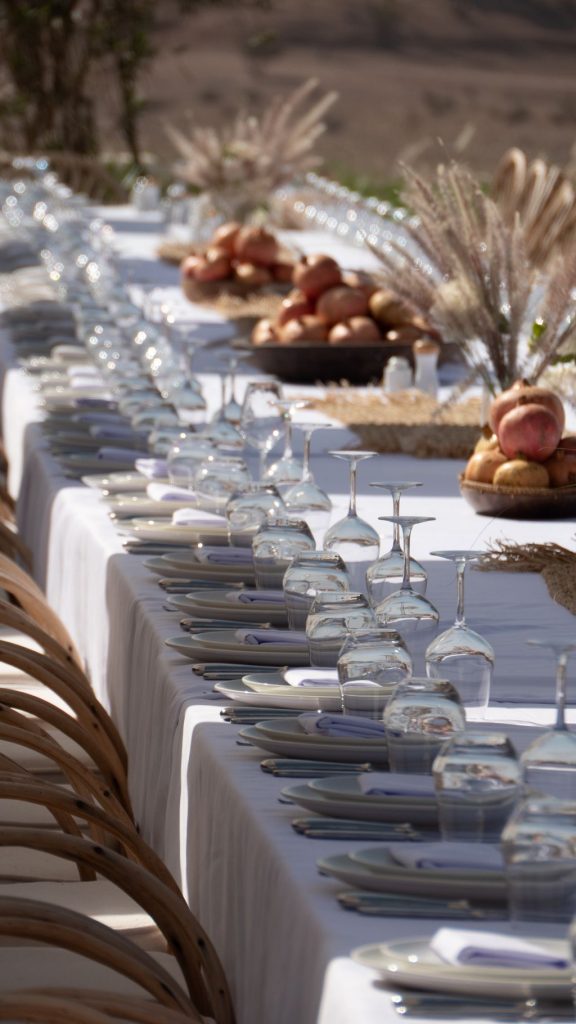 Long banquet table set with white linens, rows of glasses, pumpkins and apples ready for a big gathering