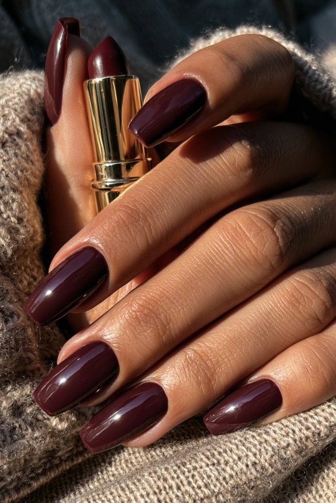 Close up of hands with long glossy plum nails holding a gold lipstick against a knit sweater