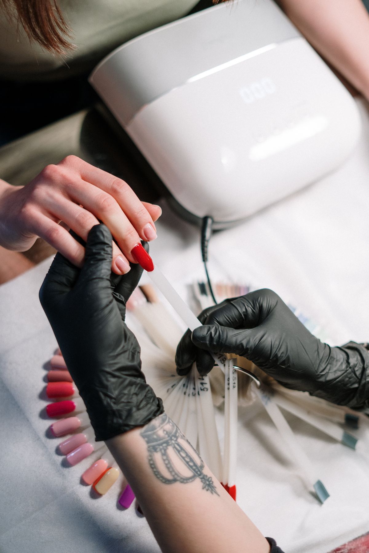 Nail technician in black gloves working on a client's hand with red polish, with a nail machine nearby
