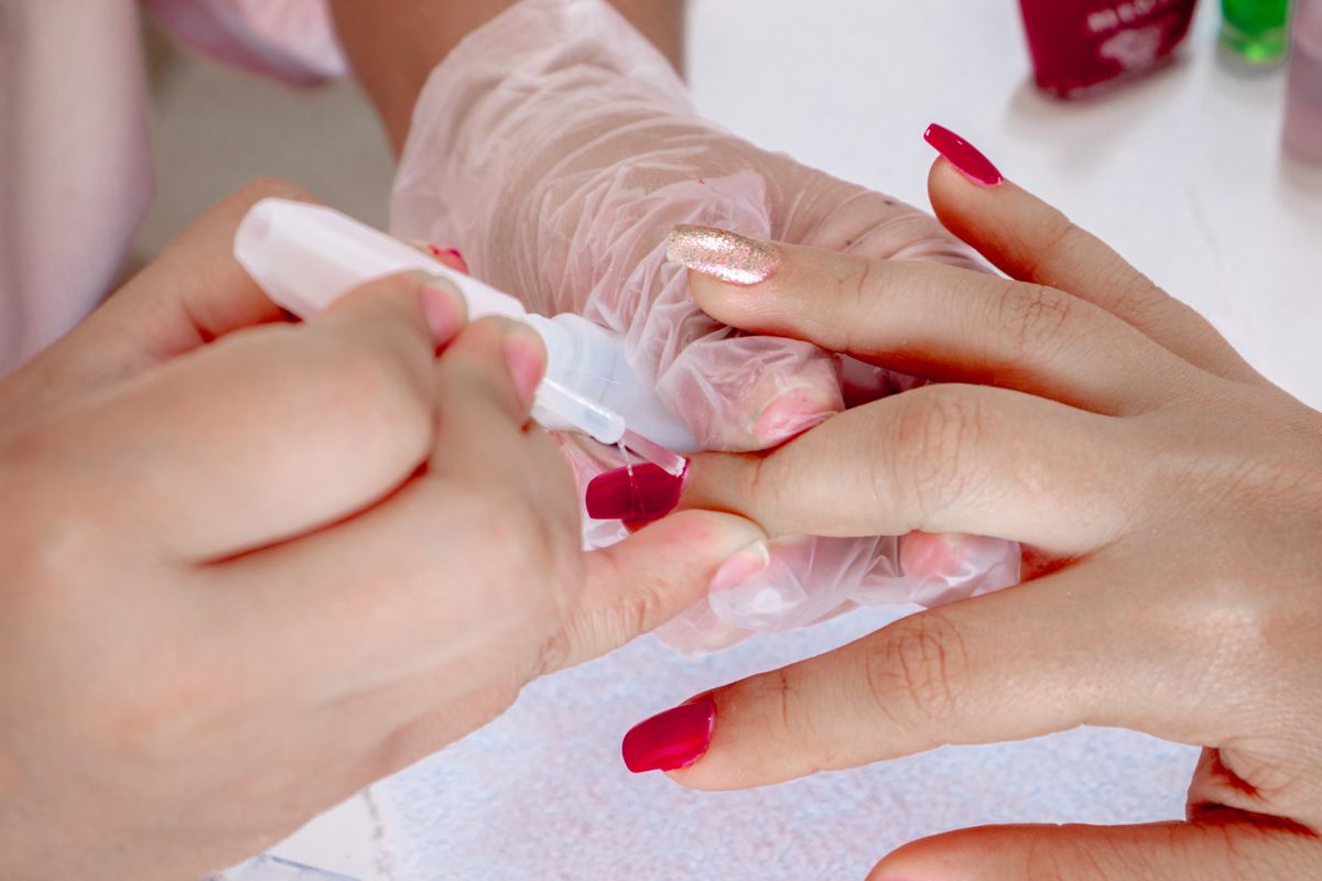 Nail technician in clear gloves using a white pen shaped tool on a client's nail with red polish