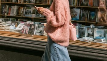 Woman in an oversized pink knit sweater with a long denim skirt and white sneakers walking past a bookstore