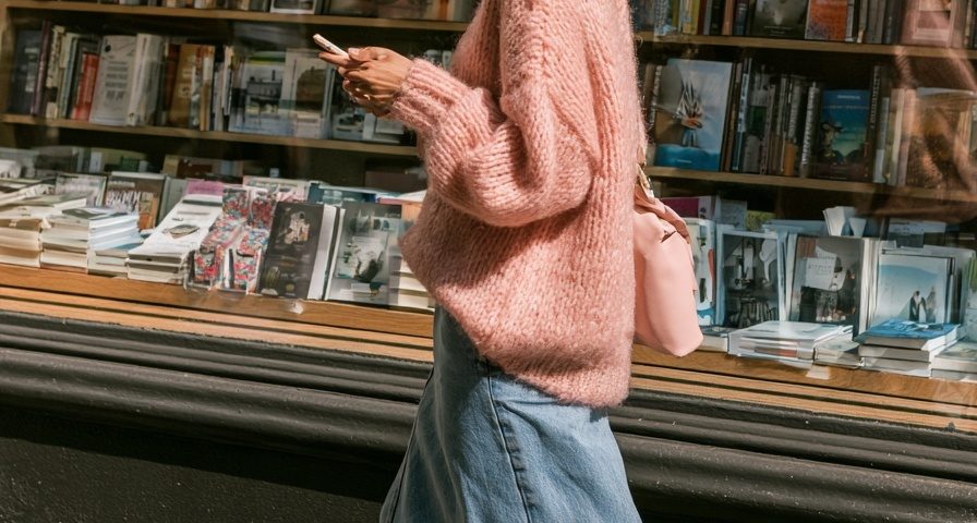 Woman in an oversized pink knit sweater with a long denim skirt and white sneakers walking past a bookstore