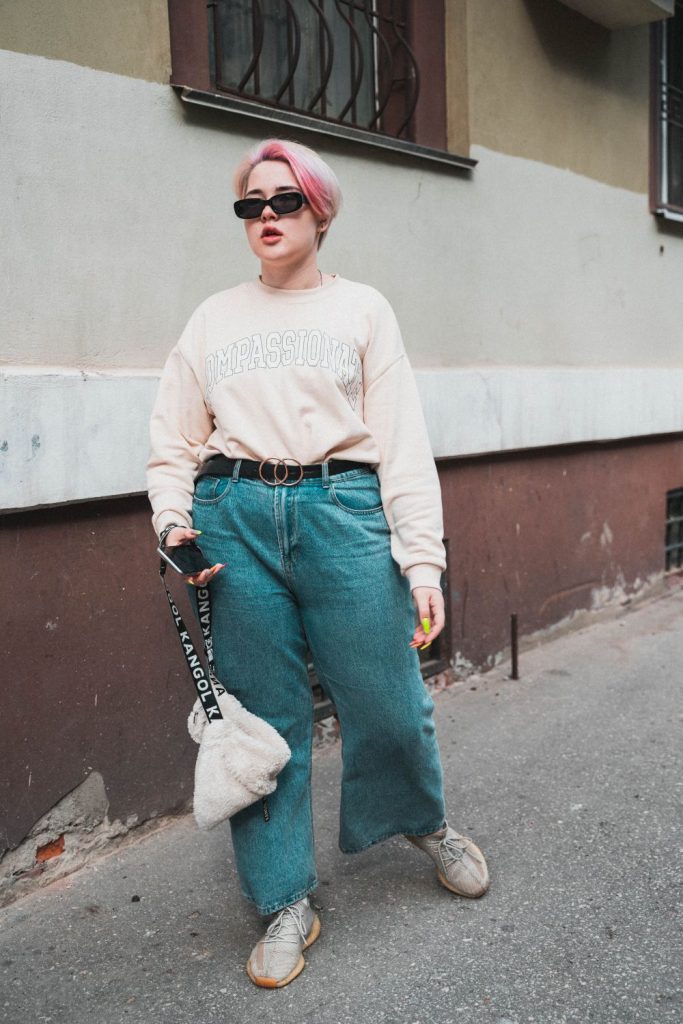 Woman in a cream oversized sweater with a black belt, wide-leg blue jeans, a small white fluffy bag and sunglasses