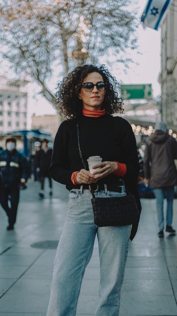 Woman with curly hair and sunglasses in a black sweater with an orange turtleneck underneath, jeans, a black quilted bag and a coffee