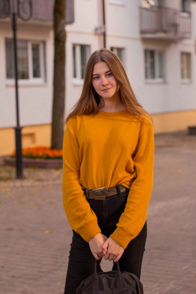 Woman in a mustard yellow oversized sweater with a dark belt and dark trousers standing on a city street