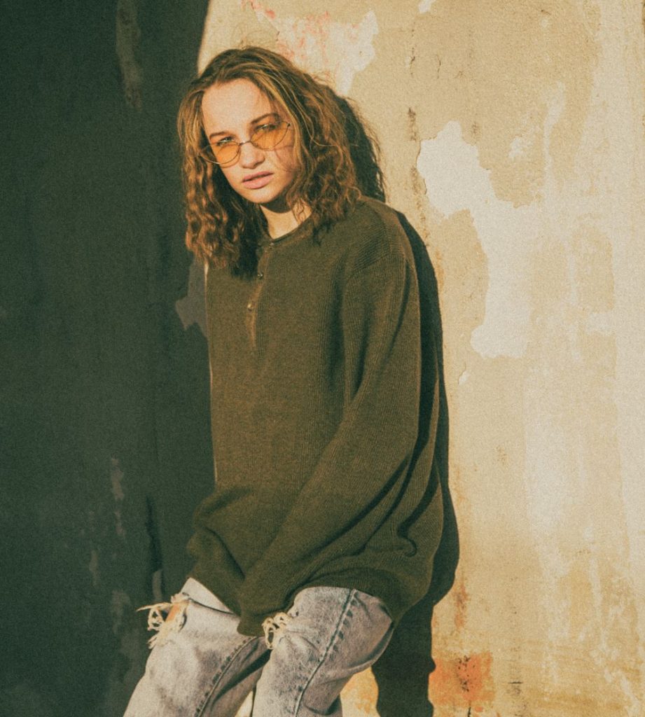 Woman with curly hair in an oversized dark olive green sweater and light jeans leaning on a textured wall