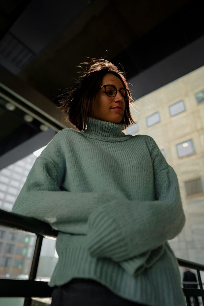 Woman in glasses wearing an oversized sage green turtleneck sweater with wide cuffs