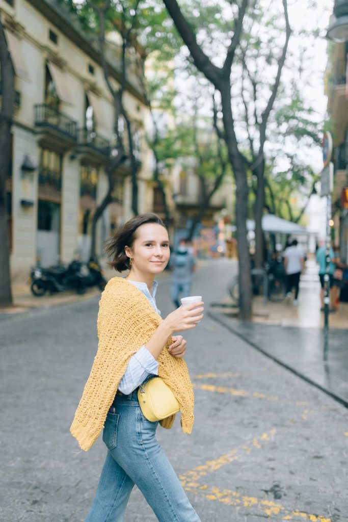 Woman on a city street with a yellow knit cardigan draped over her shoulders, a white shirt, jeans, a small yellow bag and a coffee cup