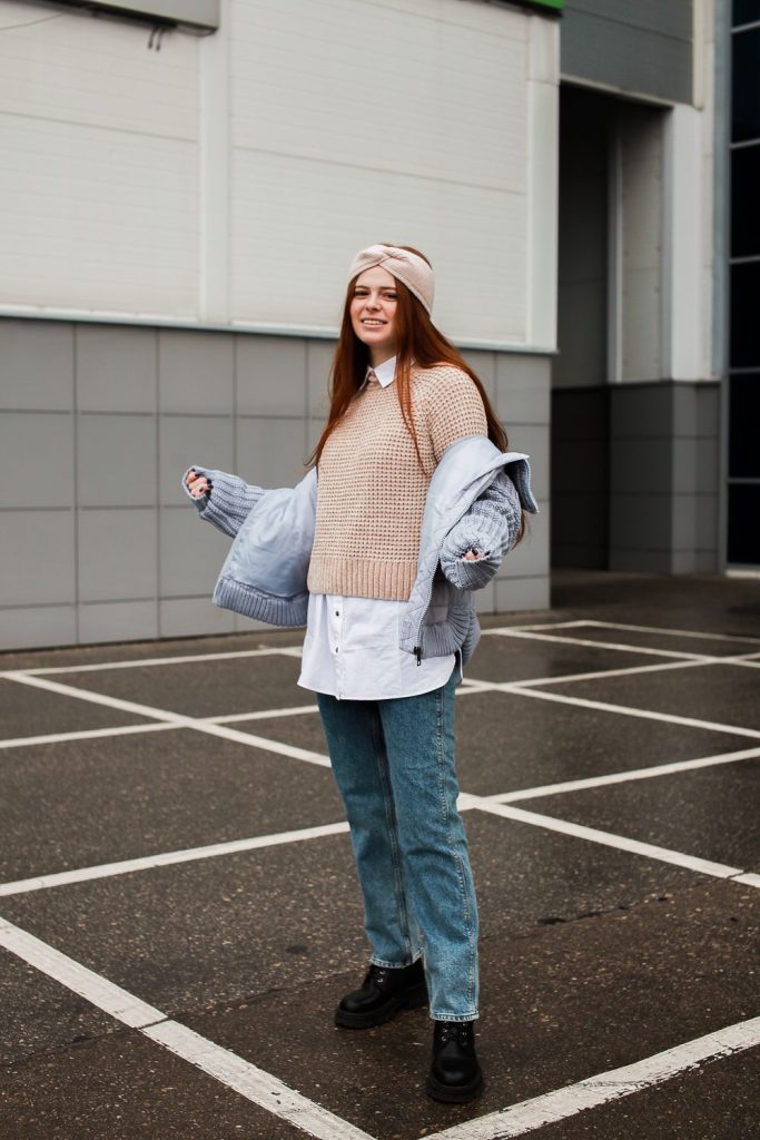 Woman in a blush knit vest over a white shirt and light jeans with a blue puffer jacket draped over her shoulders and a headband