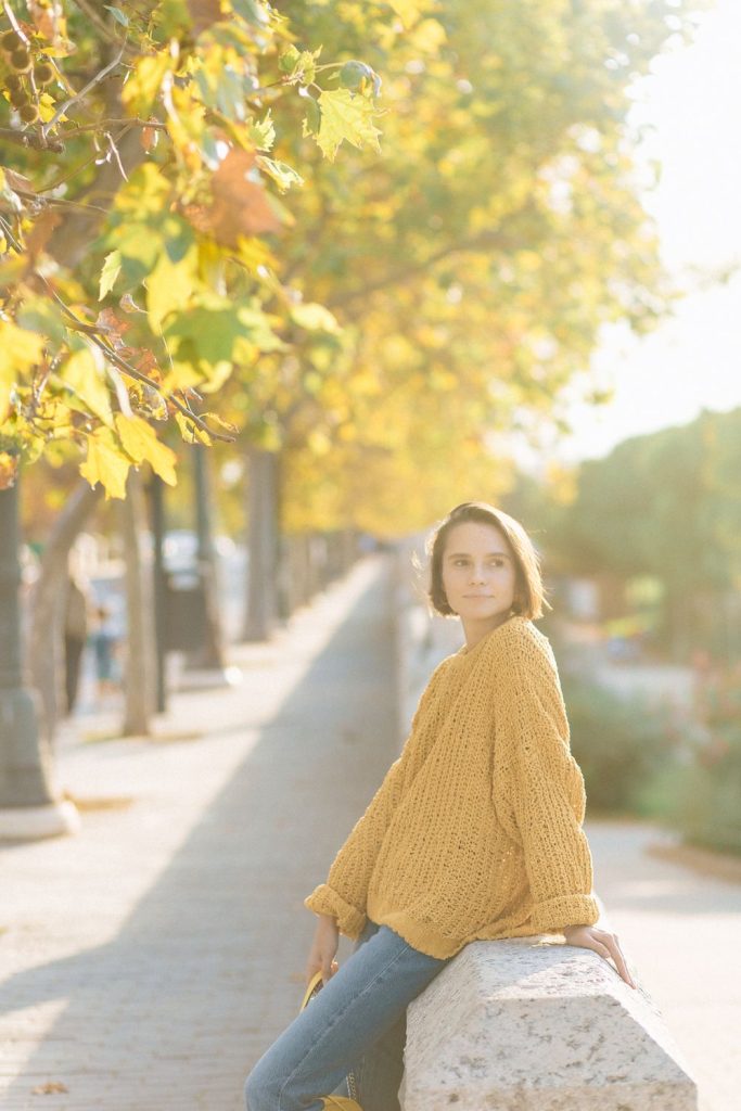Woman sitting outdoors in a chunky yellow knit sweater with golden autumn trees behind her