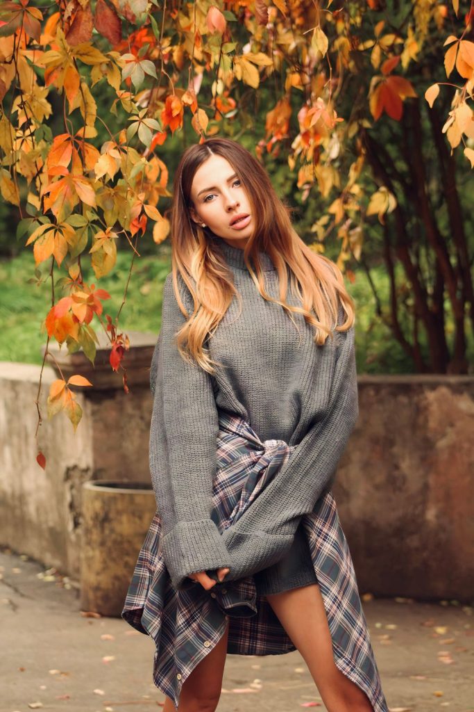 Woman in an oversized grey sweater with a plaid shirt tied around her waist standing in front of autumn leaves