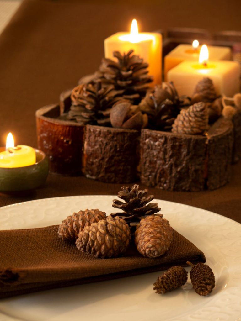 Pillar candles glowing behind a wooden bowl of pinecones, with more pinecones on a plate for a rustic fall centerpiece