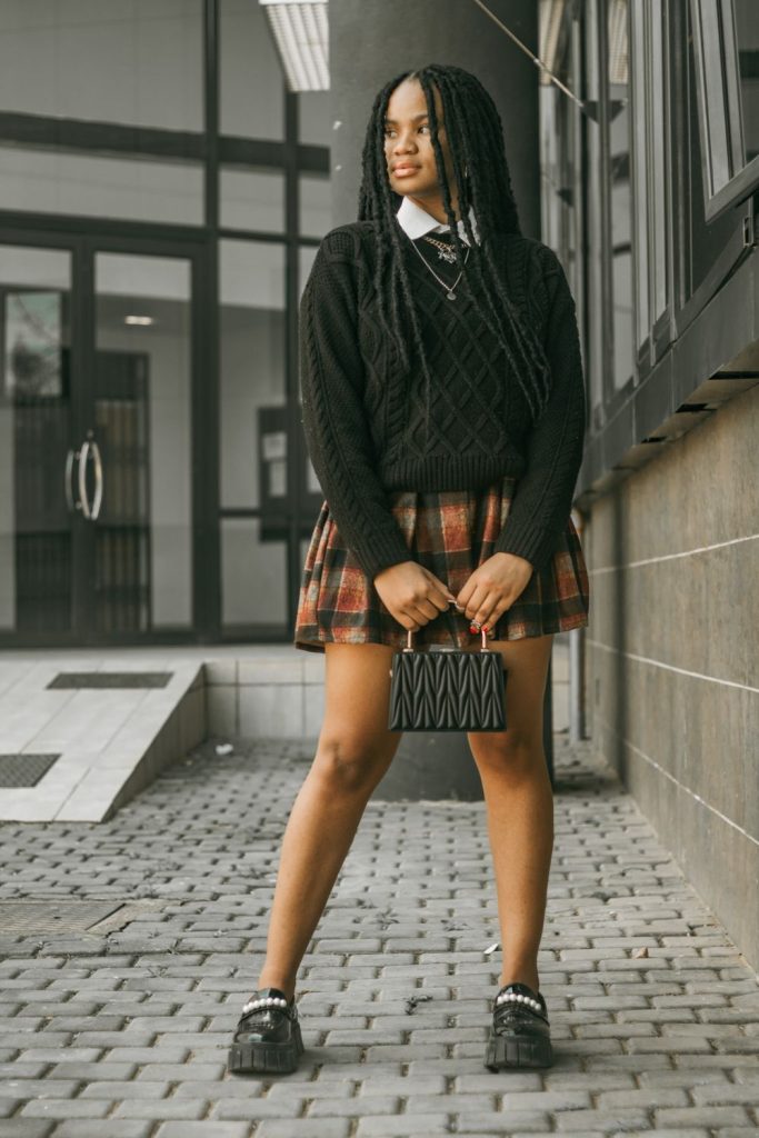 Woman in a black sweater with a white collar showing, an orange and brown plaid mini skirt, a small black bag and black shoes
