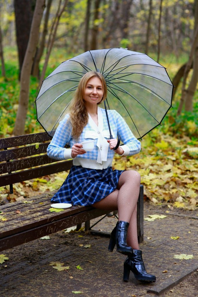 Woman on a park bench in a light blue cardigan with a bow detail and a blue plaid skirt with black boots and a clear umbrella