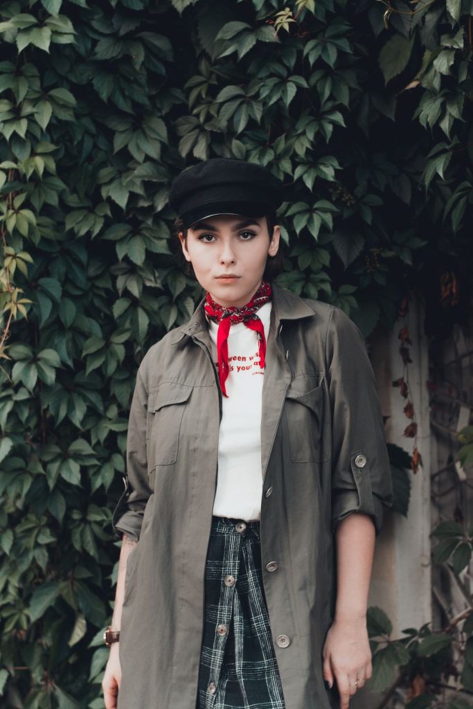 Woman in an olive utility jacket, a white top, a red neckerchief and a flat cap with a plaid skirt standing in front of ivy
