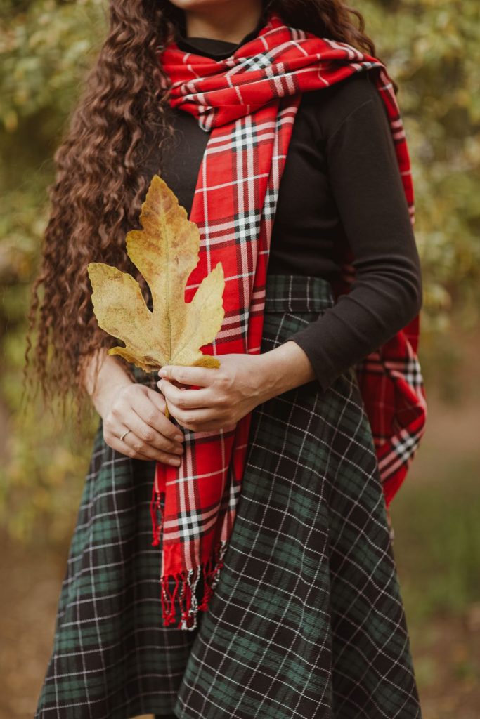 Woman in a red plaid scarf, a black long-sleeve top and a dark green plaid skirt holding a golden leaf