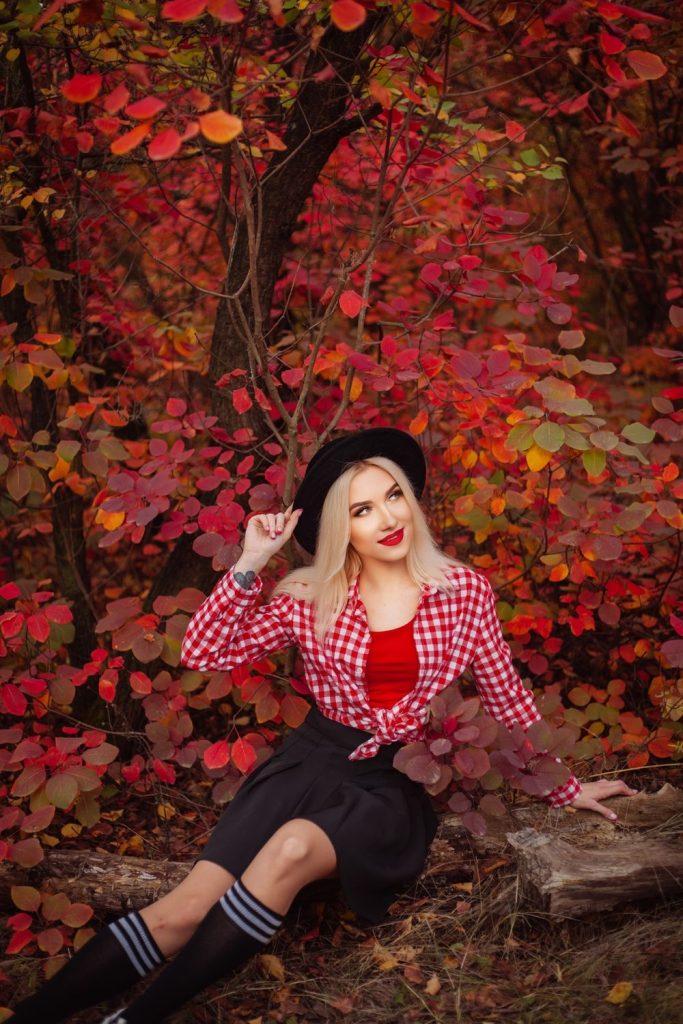 Woman in a red and white checked shirt tied over a red top with a black skirt, striped socks and a black hat among red autumn leaves