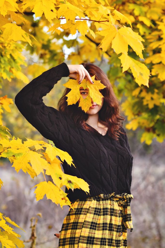 Woman in a black cable knit sweater and a yellow and black plaid skirt holding a yellow maple leaf in front of her face