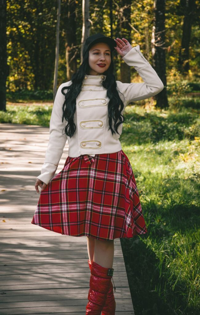 Woman in a cream button-front jacket with a full red plaid skirt and red boots walking along a park path