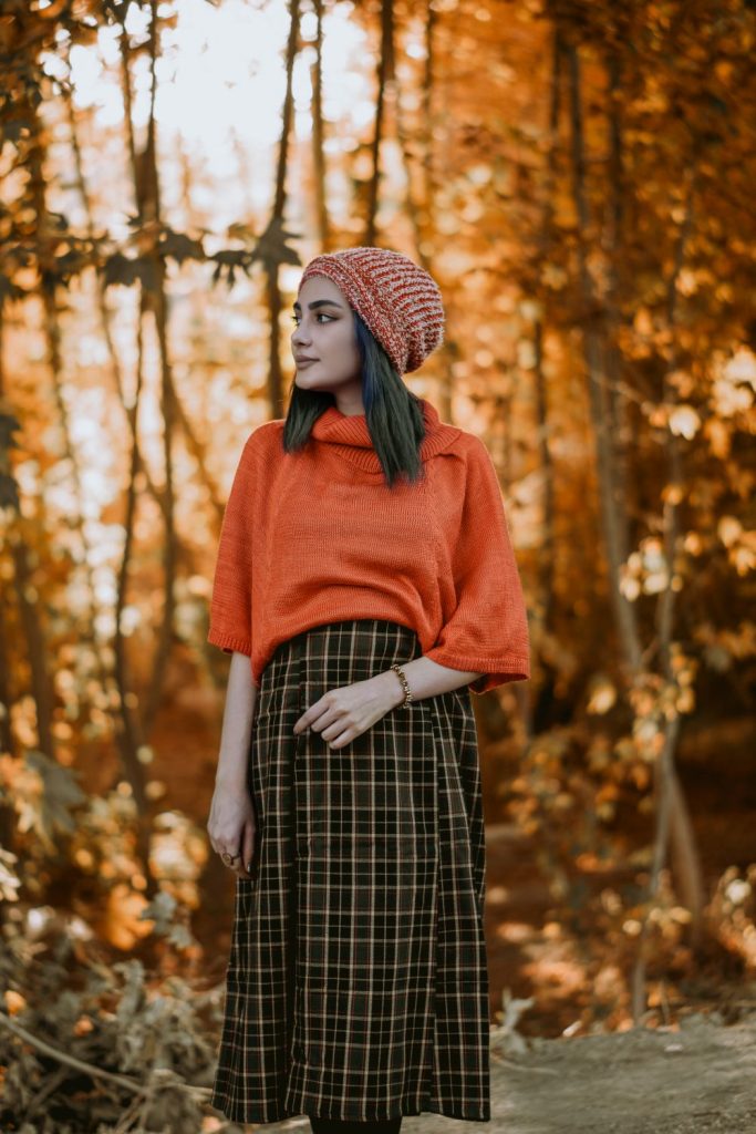 Woman in an orange knit top, a knit beanie and a long dark green and black plaid skirt standing in an orange autumn forest