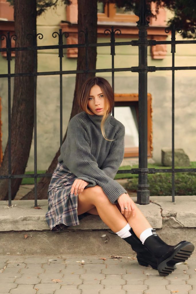 Woman sitting on a step in a grey sweater, a grey plaid skirt, white socks and black chunky boots in front of an iron fence