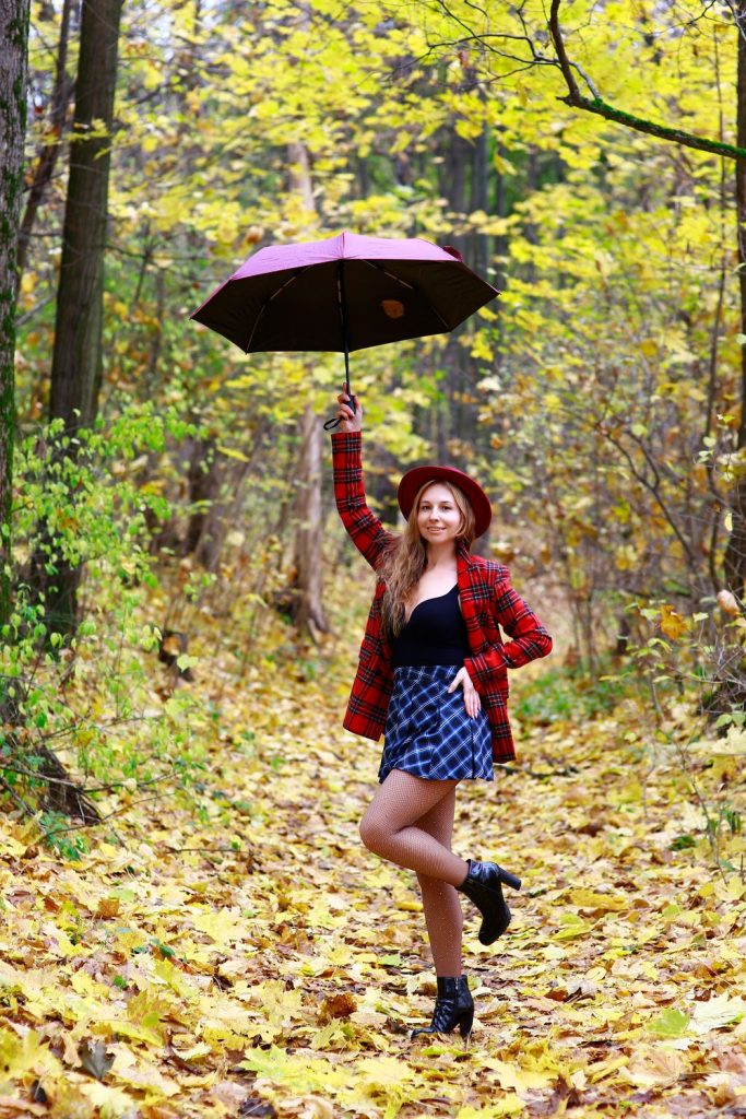 Woman in a red plaid jacket and a blue plaid skirt with a red hat, black boots and a pink umbrella among yellow leaves