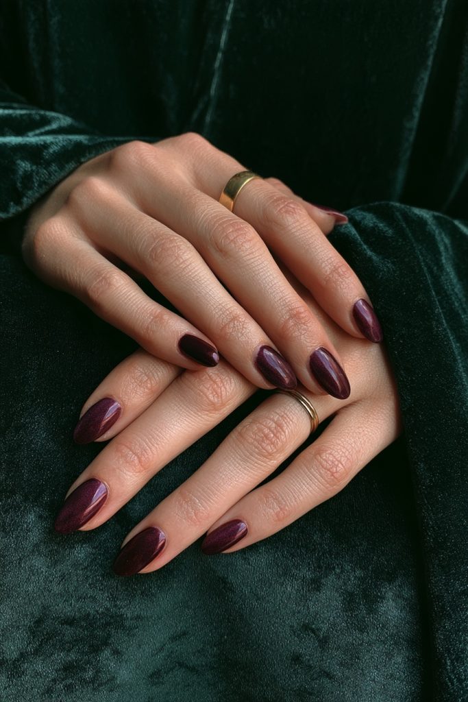 Close up of hands with glossy plum nails and a thin ring against dark green velvet