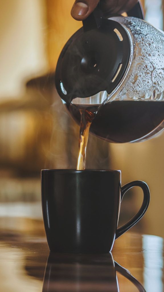 Hand pouring hot coffee from a glass carafe into a black mug at a simple self-serve coffee spot