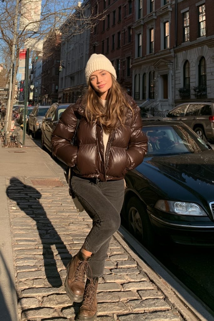 Woman in a chocolate brown puffer jacket and cream knit beanie with dark jeans and brown boots on a sunny street