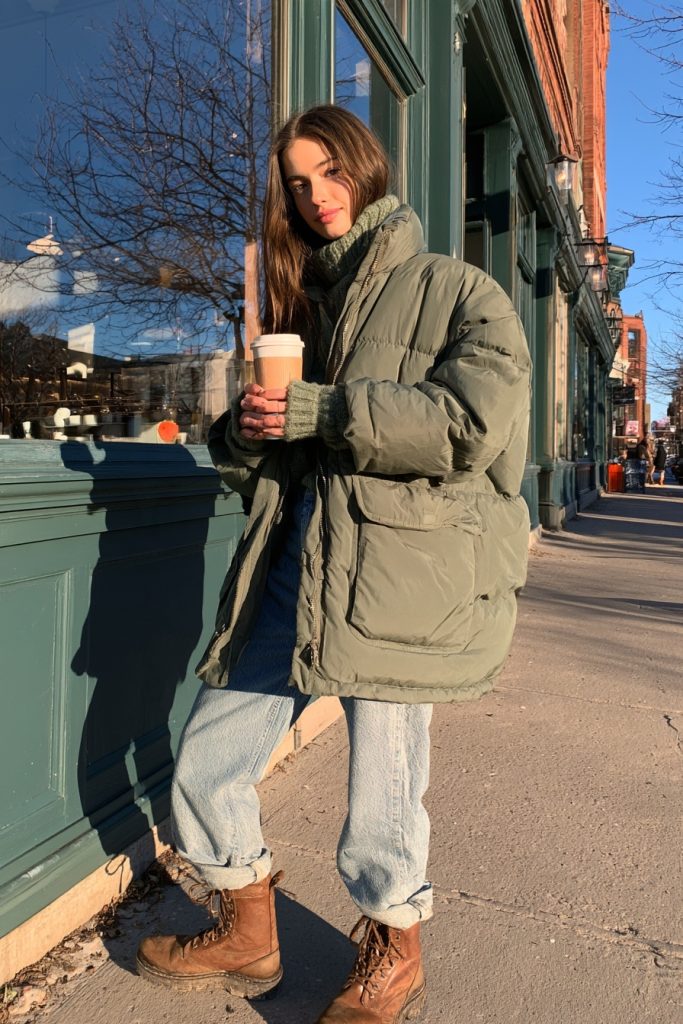 Woman in an oversized olive green puffer jacket with light straight jeans and brown boots holding a takeaway coffee cup